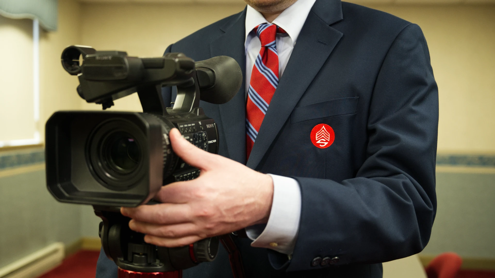 A close shot of a legal videographer wearing a suit jacket that has the Sargents S logo and holding his camera on a tripod, at chest height.